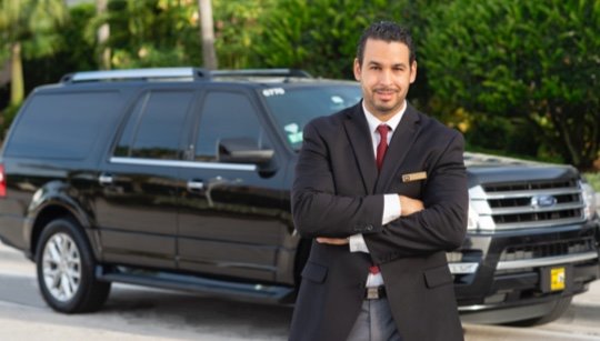 Happy, smiling male luxury chauffeur stands beside a black Mears SUV