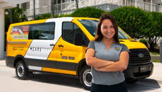 Happy, smiling female taxi driver stands beside a white and yellow Mears Taxi van outside the Orlando Convention Center