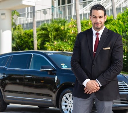 Handsome, smiling luxury private car service chauffeur in black jacket, white shirt, maroon tie and grey pants stands beside a black Mears luxury sedan