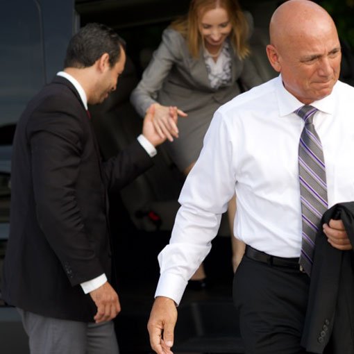 A chauffeur assists a woman in grey suit to exit a Mears Luxury Van while another gentleman passenger already exited the vehicle and is walking away